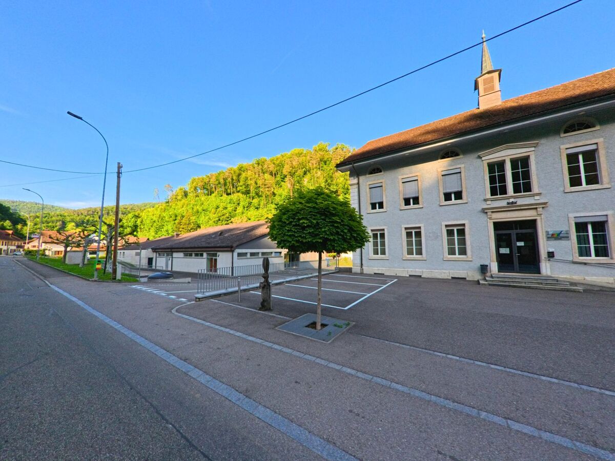 Das Schulhaus von Soyhières (2805, Kanton Jura) mit kleinem Dachreiter liegt an einer breiten Strasse, umgeben von Parkflächen und grüner Hangkulisse.