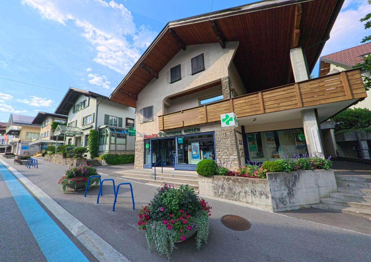 In Spiez (3700, Kanton Bern) liegt eine Apotheke mit Blumenbepflanzung an einer leicht ansteigenden Strasse, eingerahmt von Wohnhäusern mit traditionellen Fassaden.