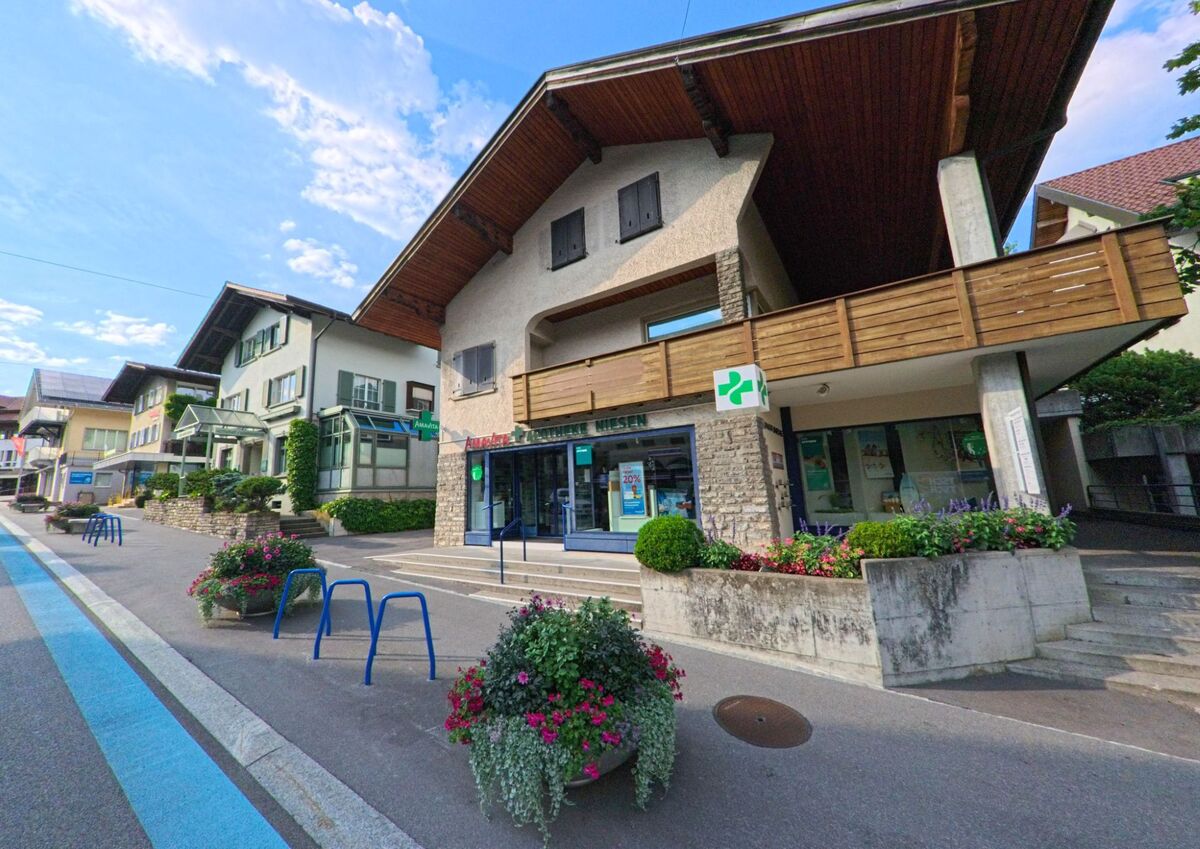 In Spiez (3700, Kanton Bern) liegt eine Apotheke mit Blumenbepflanzung an einer leicht ansteigenden Strasse, eingerahmt von Wohnhäusern mit traditionellen Fassaden.