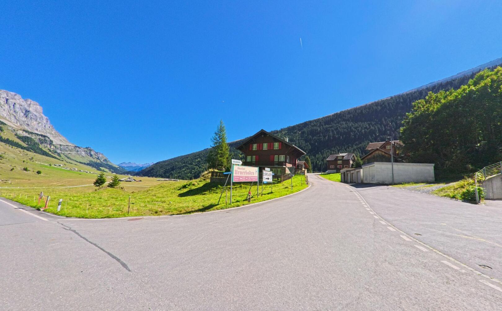 Am Ortsrand von Spiringen (8751, Kanton Uri) fuehrt eine Strasse durch eine weite, sonnige Talschaft mit vereinzelten Haeusern und gruenen Wiesen.