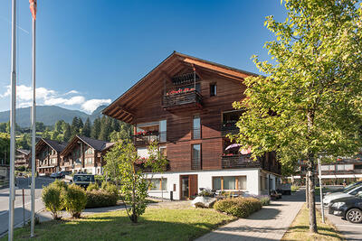 In Grindelwald (3818, Kanton Bern) steht ein modernes Holzhaus mit Balkonen und Gartenanlage vor einer alpinen Bergkulisse bei sonnigem Wetter.