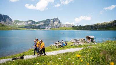 In Kerns (6064, Kanton Obwalden) spazieren Menschen mit Hund entlang des Melchsees, waehrend im Hintergrund die Berge und Gebaeude der Sportbahnen Melchsee-Frutt zu sehen sind.