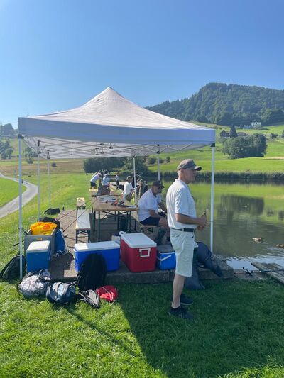 In Wollerau (8807, Kanton Schwyz) angeln Mitglieder des Sportfischerverein Hoefe unter einem Sonnenschutzdach an einem ruhigen Gewaesser inmitten gruener Huegel.