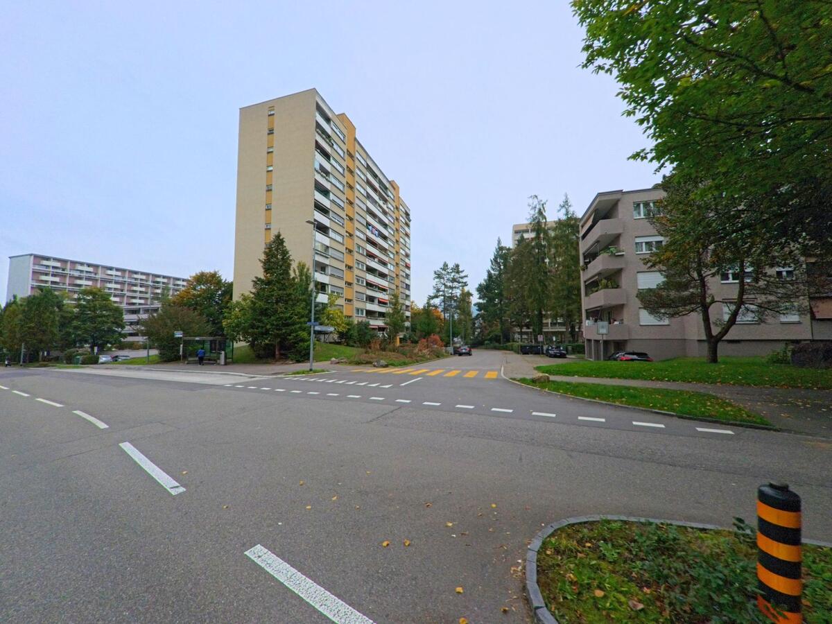 In Spreitenbach (8957, Kanton Aargau) praegt ein hohes Wohnhaus mit Balkonen die Kreuzung, waehrend weitere Mehrfamilienhaeuser und Baeume die Umgebung bestimmen.