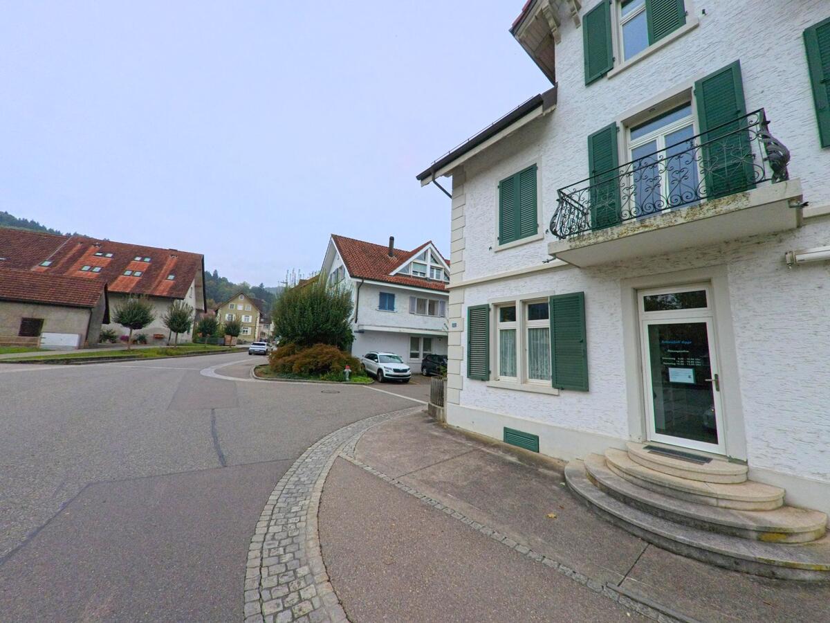In einer ruhigen Wohnstrasse von Spreitenbach (8957, Kanton Aargau) steht ein Haus mit grünen Fensterlaeden und kleinem Balkon unmittelbar an einer sanft geschwungenen Kurve.