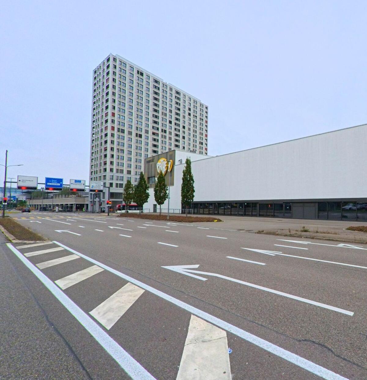 Eine breite mehrspurige Strasse in Spreitenbach (8957, Kanton Aargau) verläuft am grossen Pathé-Gebaeude entlang, vor dem ein markantes Hochhaus den urbanen Bereich praegt.