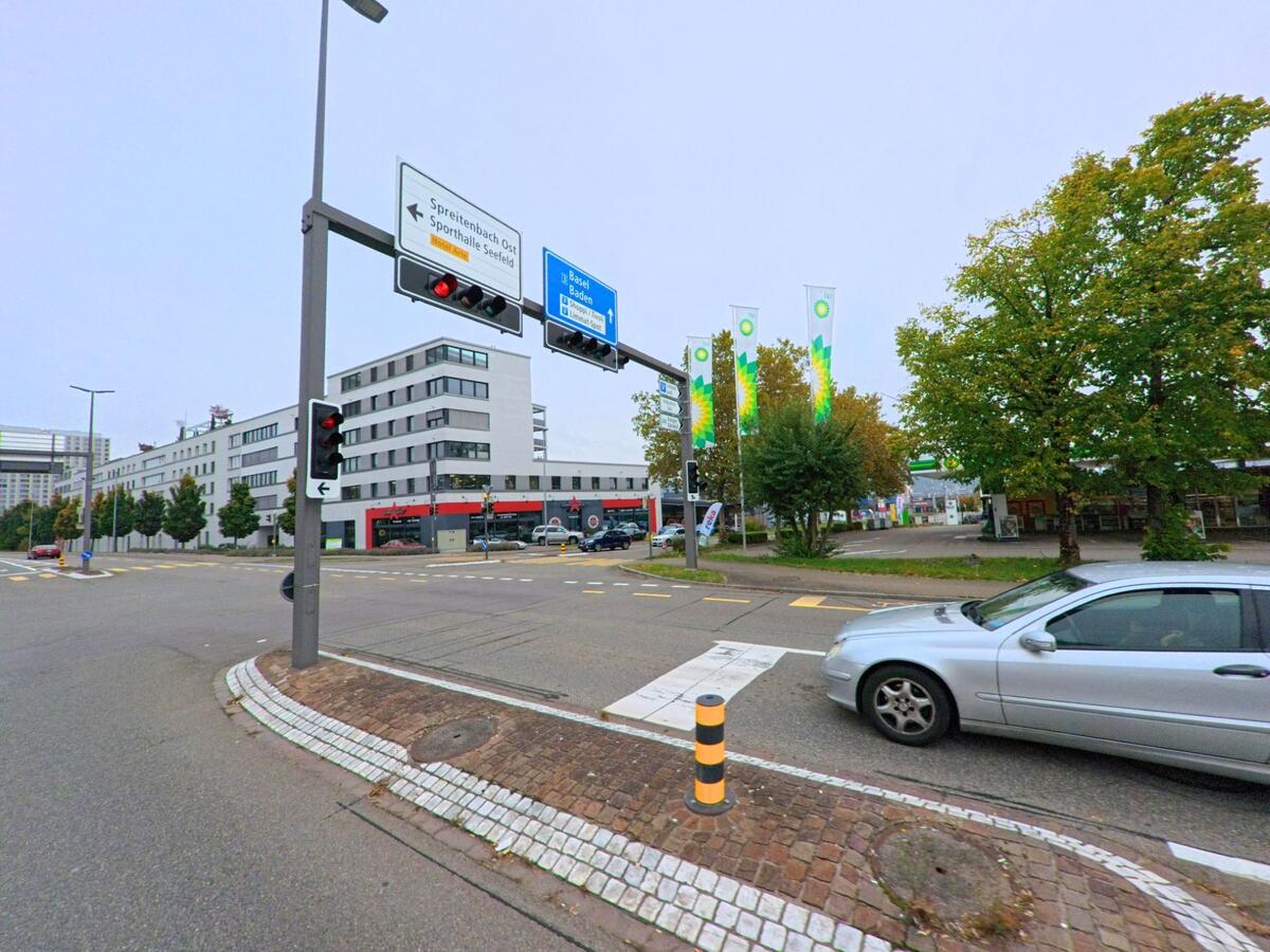 An einer stark befahrenen Kreuzung in Spreitenbach (8957, Kanton Aargau) stehen moderne Wohn- und Gewerbegebaeude, waehrend auf der Gegenseite eine Tankstelle mit hohen Fahnen sichtbar ist.