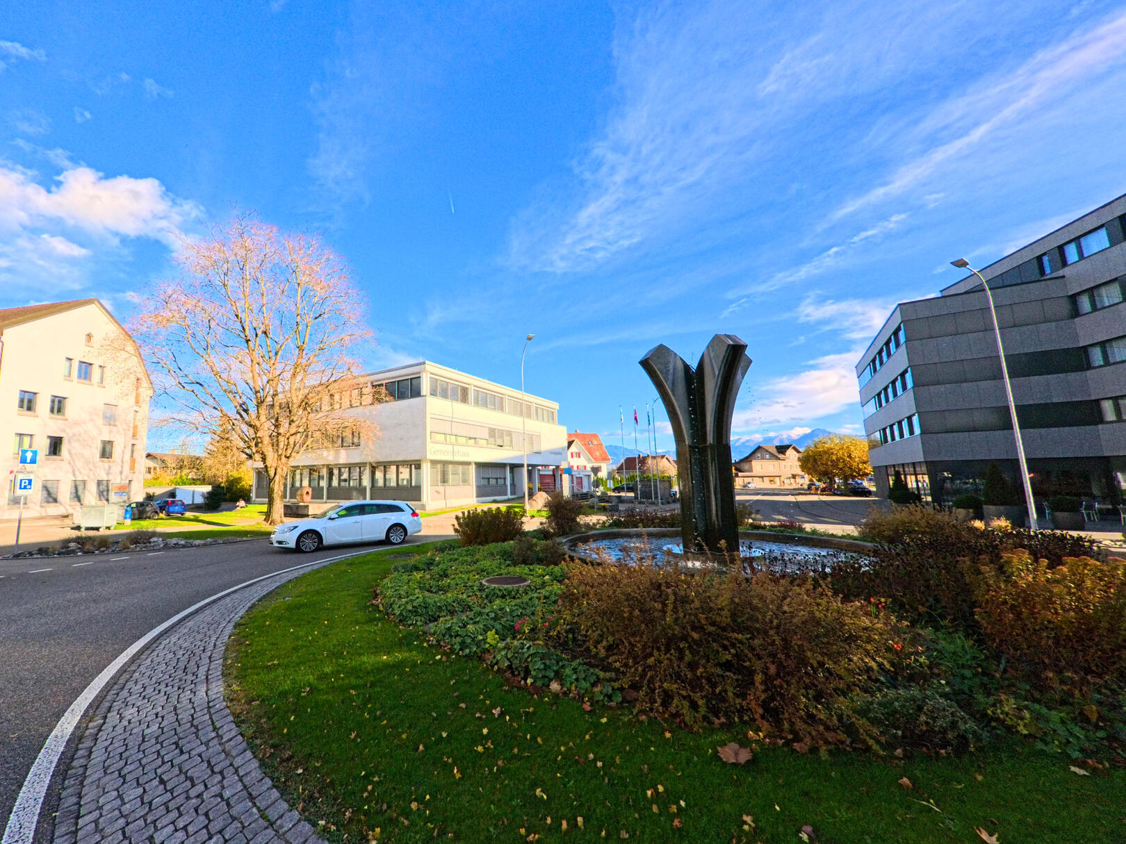 Ein Kreisverkehr mit Brunnenplastik und Verwaltungsbauten praegt das Ortszentrum von St. Margarethen (9430, Kanton St. Gallen) unter klarem Himmel.