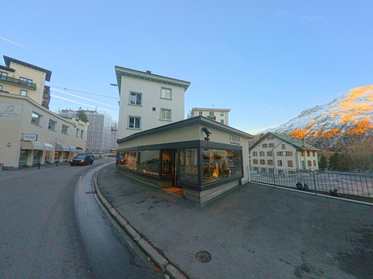 An einer sanften Kurve steht ein Eckgebäude mit grossen Schaufenstern in St. Moritz (7500, Kanton Graubünden), während dahinter Wohnhäuser und ein beleuchteter Berghang sichtbar sind.