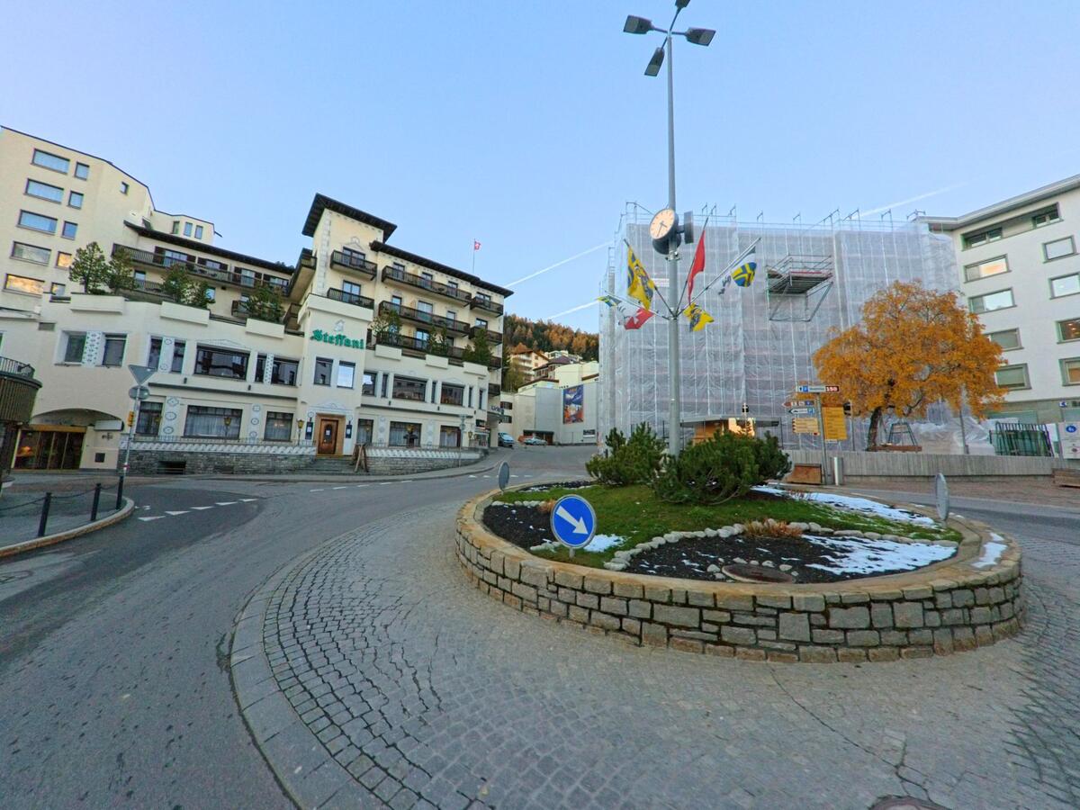 An einem Kreisverkehr mit Uhr, Fahnen und bepflanzter Mittelinsel treffen sich mehrere Strassen in St. Moritz (7500, Kanton Graubünden) vor markanten Hotel- und Geschäftsgebäuden.