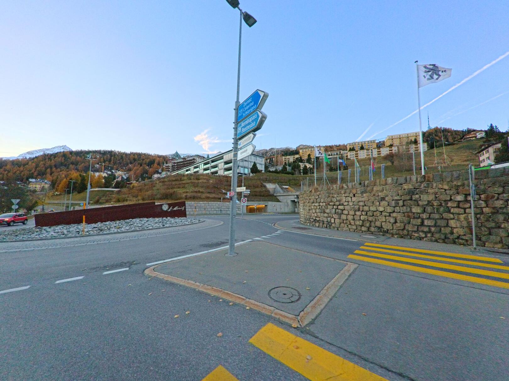 An einem Verkehrsknotenpunkt mit Wegweisern und Fussgängerstreifen öffnet sich in St. Moritz (7500, Kanton Graubünden) der Blick auf Hanglagen mit Gebäuden und Flaggenanlagen.