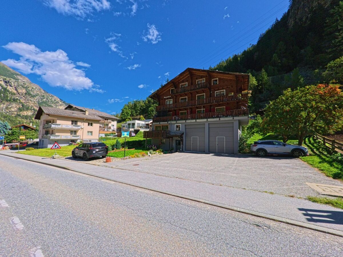 Ein dreistöckiges Holzhaus mit Balkonen steht in St. Niklaus (3924, Kanton Wallis) neben einer Strasse, umgeben von Wohnhäusern und einem Garten mit Bäumen.