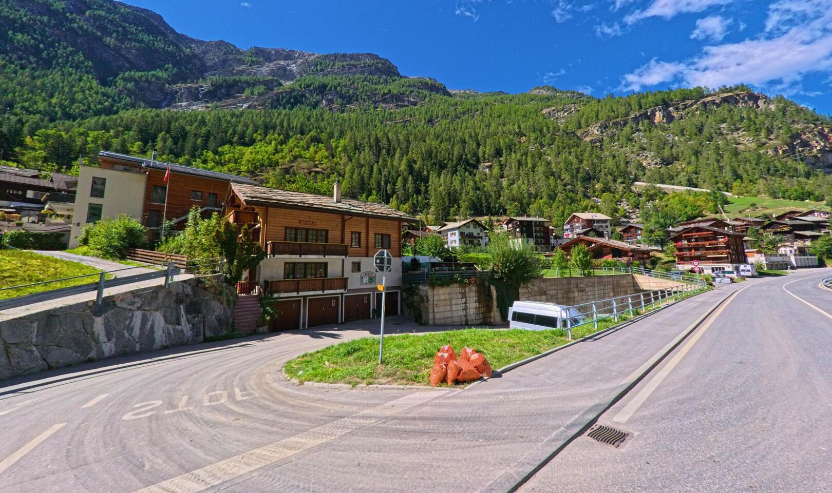 Am Rand der Strasse in St. Niklaus (3924, Kanton Wallis) stehen Holzhäuser vor einem bewaldeten Hang mit felsigem Gipfel im Hintergrund.