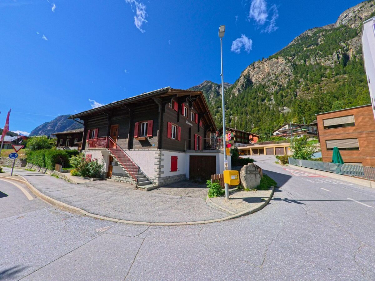 Ein traditionelles Haus mit roten Fensterlaeden und Steinsockel befindet sich in St. Niklaus (3924, Kanton Wallis) an einer Strassenecke mit Blick auf bewaldete Berge.