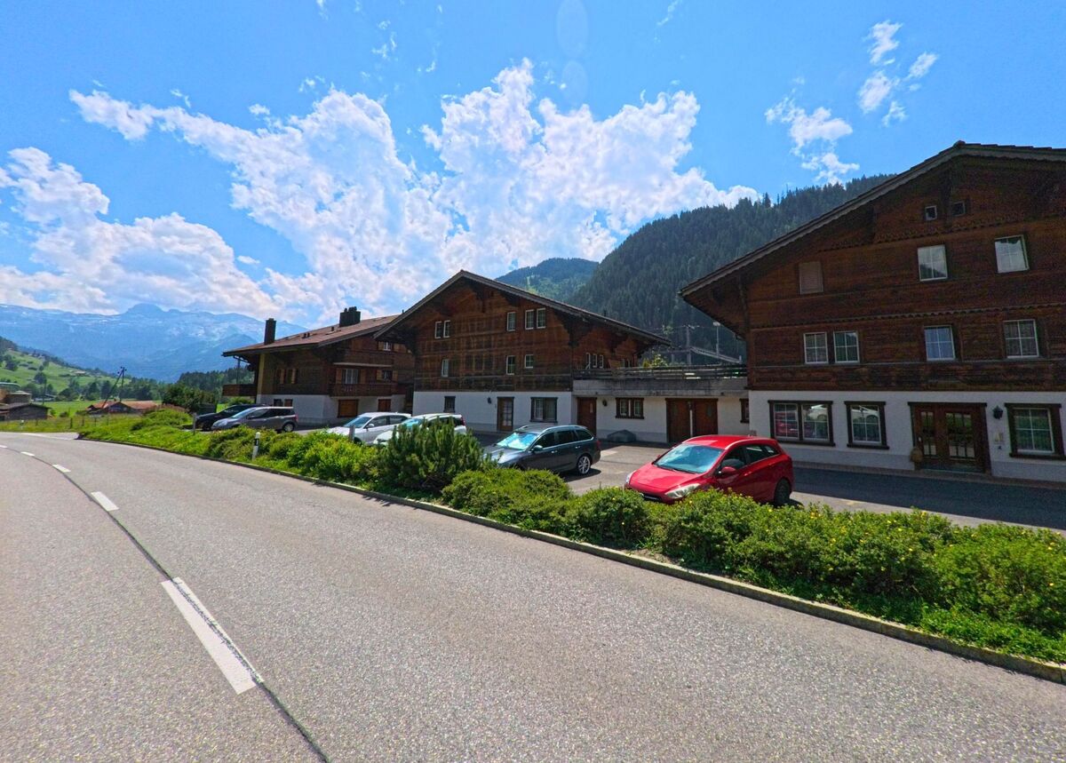 Entlang der Hauptstrasse in St. Stephan (3773, Kanton Bern) reihen sich mehrere Holzhäuser mit vorgelagerten Parkplätzen vor dem Panorama der Alpen.