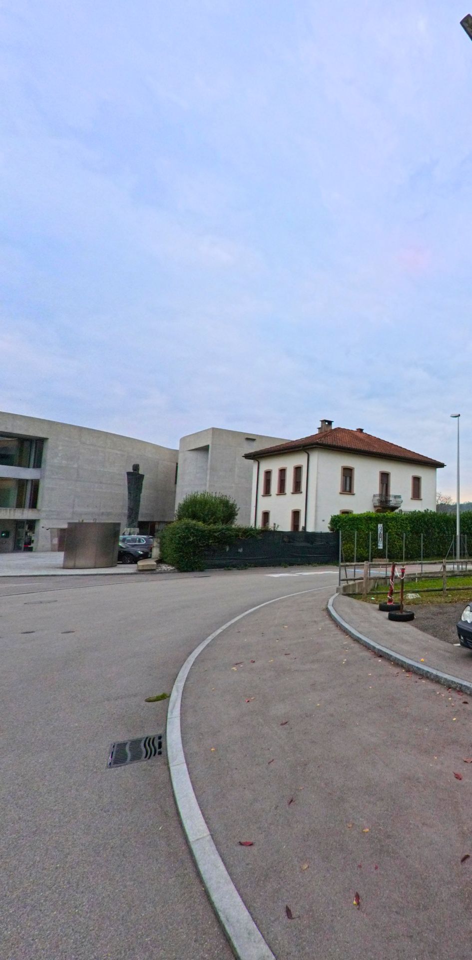 In Stabio (6854, Kanton Tessin) treffen moderne Betonarchitektur und ein traditionelles Wohnhaus mit rotem Ziegeldach aufeinander, getrennt durch eine schmale Strassenkurve.