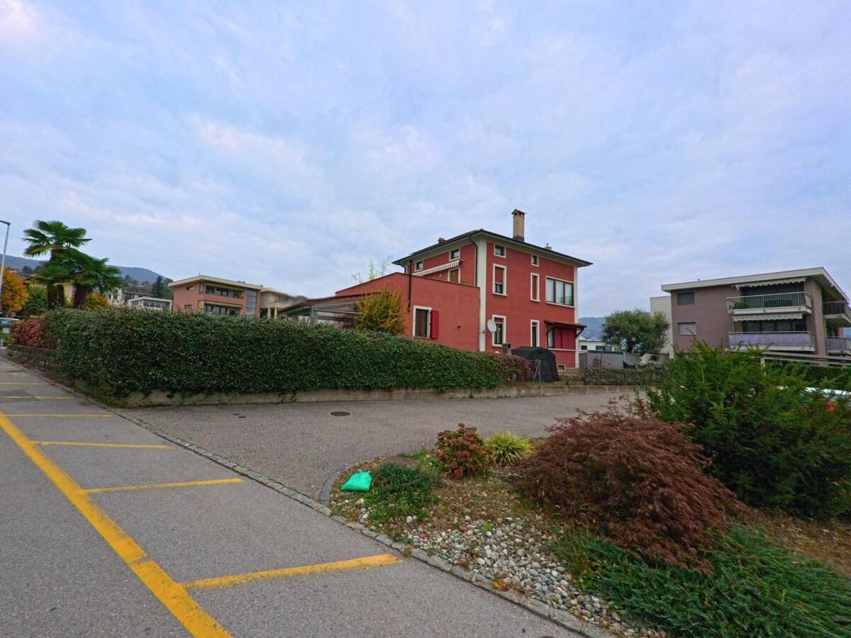 Ein rotes Wohnhaus mit Garten und umliegenden modernen Gebäuden steht in Stabio (6854, Kanton Tessin) an einer ruhigen Strasse mit Parkstreifen.