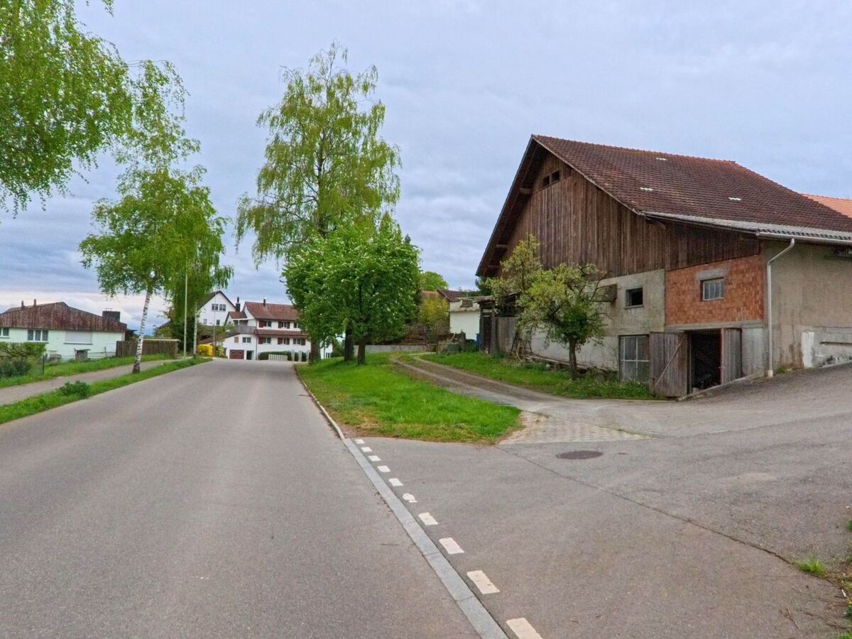 An einer leicht ansteigenden Strasse in Stadel bei Niederglatt (8174, Kanton Zürich) liegt ein grosser Bauernhof mit Nebengebaeuden, flankiert von Baeumen und Wohnhaeusern.