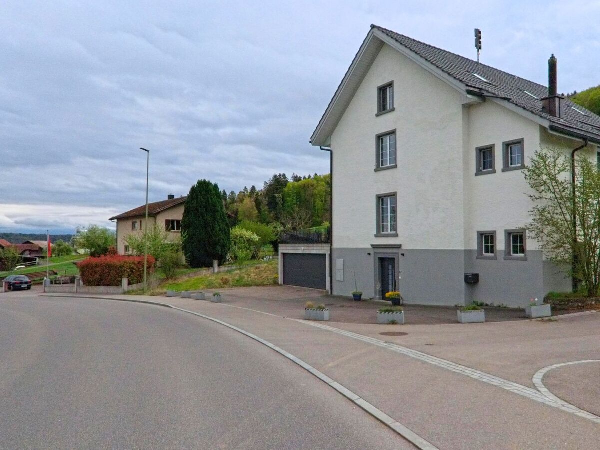 Ein mehrstöckiges Wohnhaus mit grauem Sockelbereich steht an einer Kurve in Stadel bei Niederglatt (8174, Kanton Zürich) vor einem Hintergrund aus Wiesen und bewaldeten Huegeln.