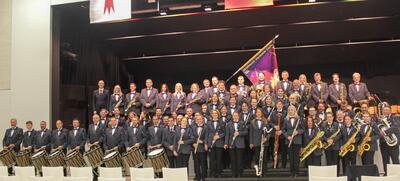 Gruppenfoto der Stadtmusik Harmonie Laufen in Laufen (4242, Kanton Baselland). Die Musikerinnen und Musiker stehen mit Instrumenten und Vereinsfahne auf einer Bühne und präsentieren das Blasorchester in einheitlicher Konzertkleidung.
