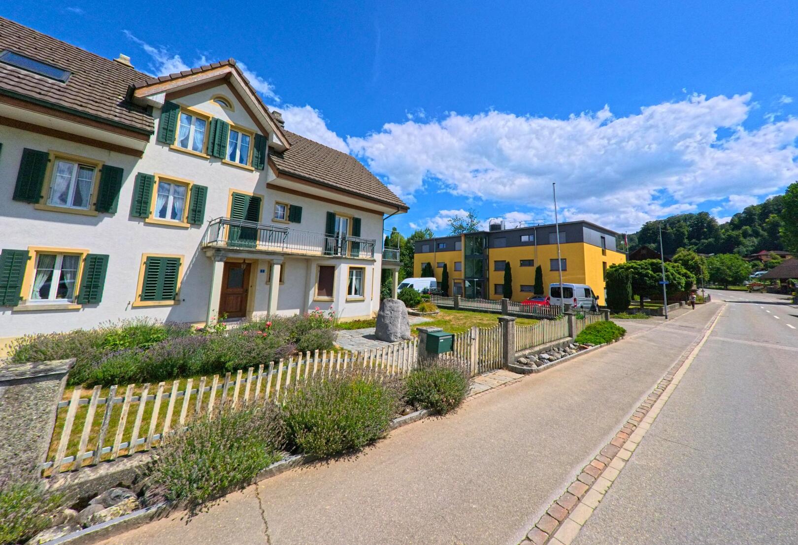 In Staffelbach (5053, Kanton Aargau) stehen ein traditionelles Wohnhaus mit gruenen Fensterlaeden und ein modernes gelbes Gebaeude nebeneinander an einer ruhig gelegenen Strasse.