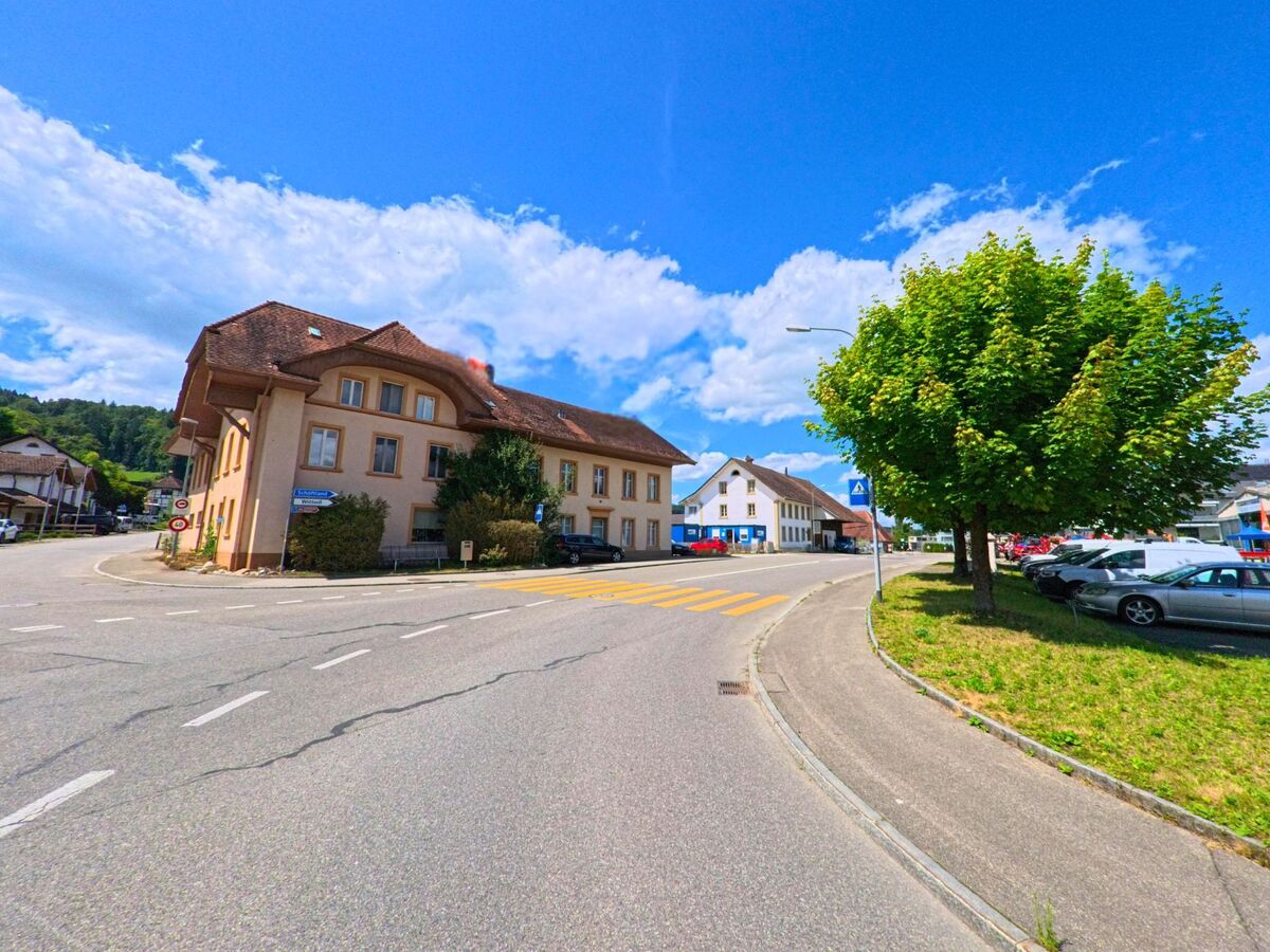 In Staffelbach (5053, Kanton Aargau) fuehrt eine Strasse mit Fussgaengerstreifen und Baumallee am Rand entlang an einem markanten Gebaeude mit Mansarddach und umliegenden Geschaeften vorbei.