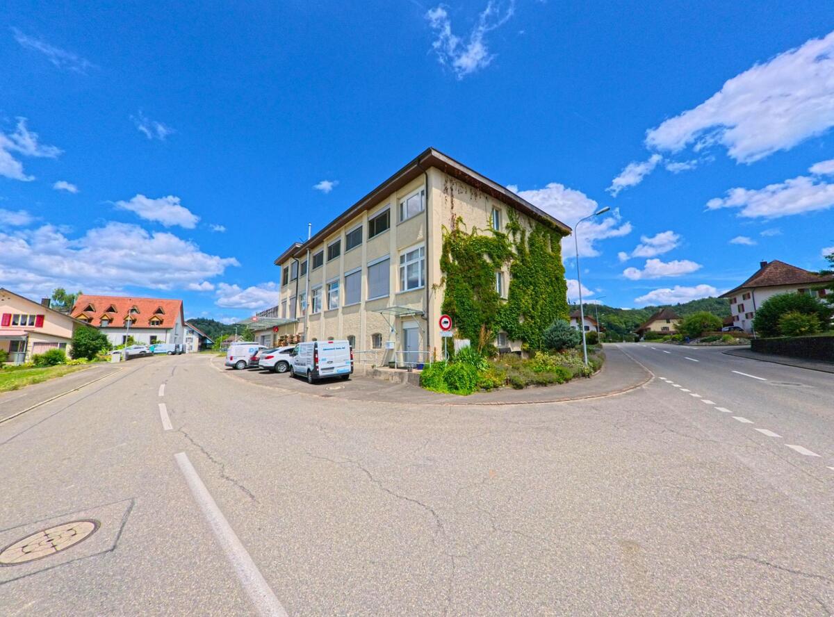 Ein von Efeu bewachsenes Gewerbegebaeude mit mehreren Lieferwagen steht an einer Strassengabelung in Staffelbach (5053, Kanton Aargau), umgeben von Wohnhaeusern und Huegellandschaft.