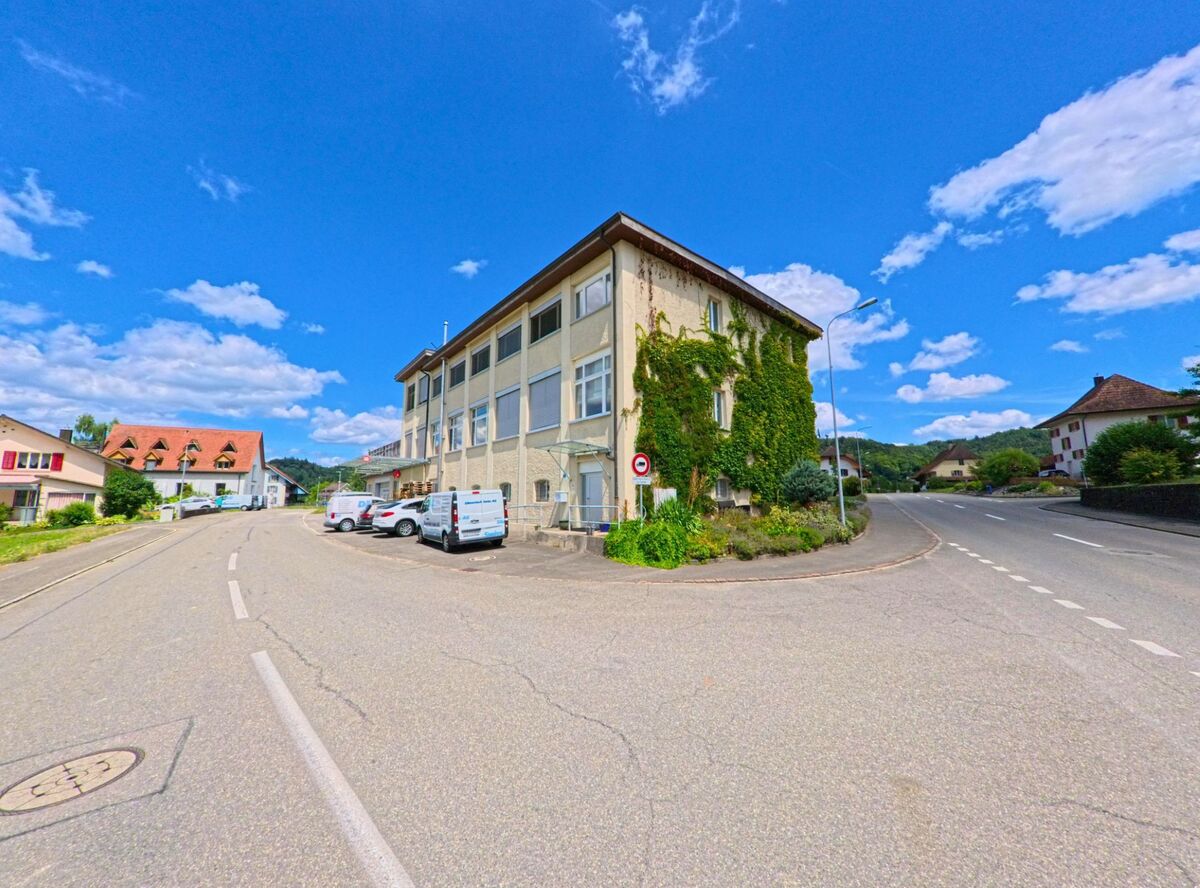 Ein von Efeu bewachsenes Gewerbegebaeude mit mehreren Lieferwagen steht an einer Strassengabelung in Staffelbach (5053, Kanton Aargau), umgeben von Wohnhaeusern und Huegellandschaft.