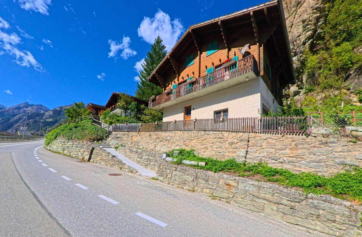 Ein traditionelles Chalet mit Holzbalkon steht in Stalden (3922, Kanton Wallis) oberhalb einer kurvigen Strasse, umgeben von Felsen und Berglandschaft.