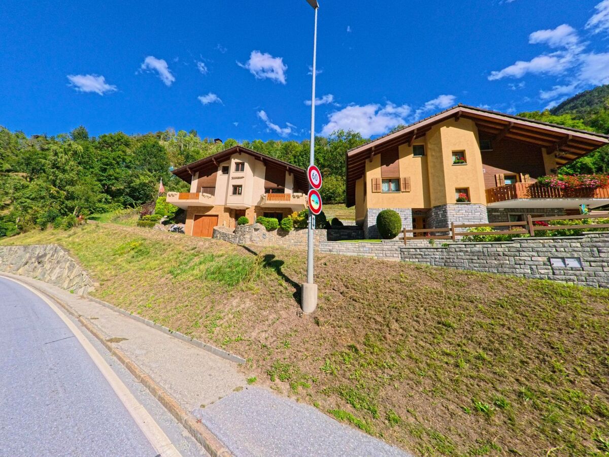 Zwei Einfamilienhaeuser mit grossen Daechern und gepflegten Gaerten stehen in Stalden (3922, Kanton Wallis) oberhalb einer Strasse am Waldrand.