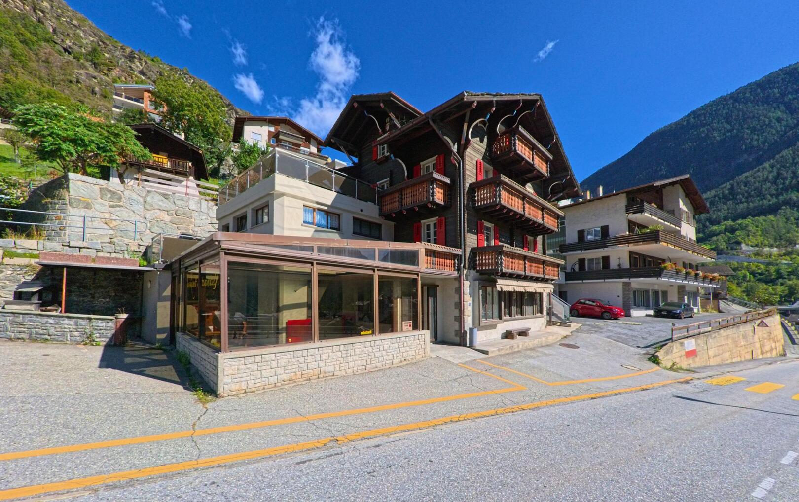 Ein traditionelles Holzhaus mit Balkonen steht in Stalden (3922, Kanton Wallis) am Hang, flankiert von modernen Wohnbauten und umgeben von steilen Berghängen.