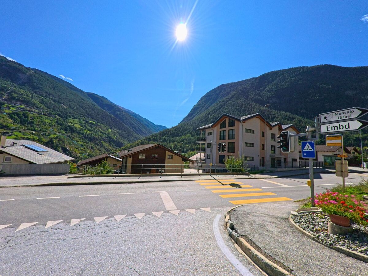 An einer Kreuzung in Stalden (3922, Kanton Wallis) stehen Wegweiser nach Moosalp, Toerbel und Embd, im Hintergrund sind moderne Wohnhaeuser und bewaldete Berghaenge sichtbar.