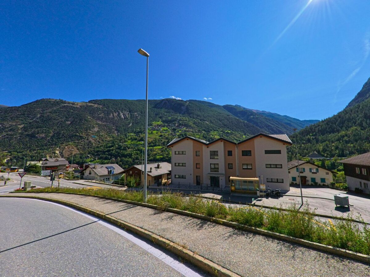 Ein groesseres modernes Gebaeude mit mehreren Wohnparteien steht in Stalden (3922, Kanton Wallis) nahe einer Strasse, umgeben von Bergen und gruener Landschaft.