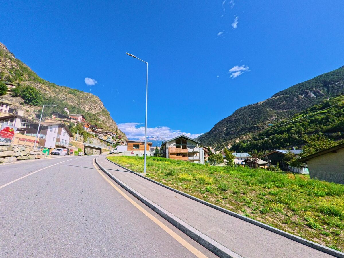 Entlang einer breiten Strasse in Stalden (3922, Kanton Wallis) stehen Wohnhaeuser am Hang, umgeben von felsigen Abhaengen und bewaldeten Bergen.