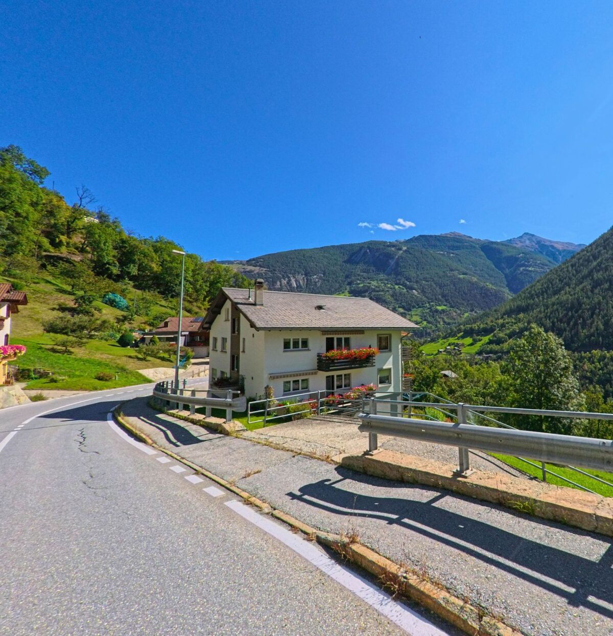 Ein helles Wohnhaus mit Blumenschmuck am Balkon steht in Stalden (3922, Kanton Wallis) an einer kurvigen Strasse mit Blick auf das gruen bewaldete Tal.