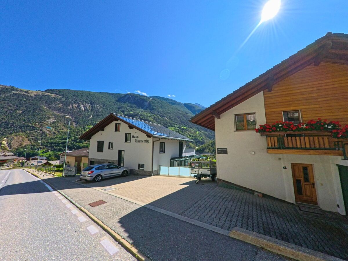 Ein helles Wohnhaus mit Solarpanelen auf dem Dach und Blumenschmuck am Balkon steht in Stalden (3922, Kanton Wallis) an einer Strasse mit Blick auf die Berge.