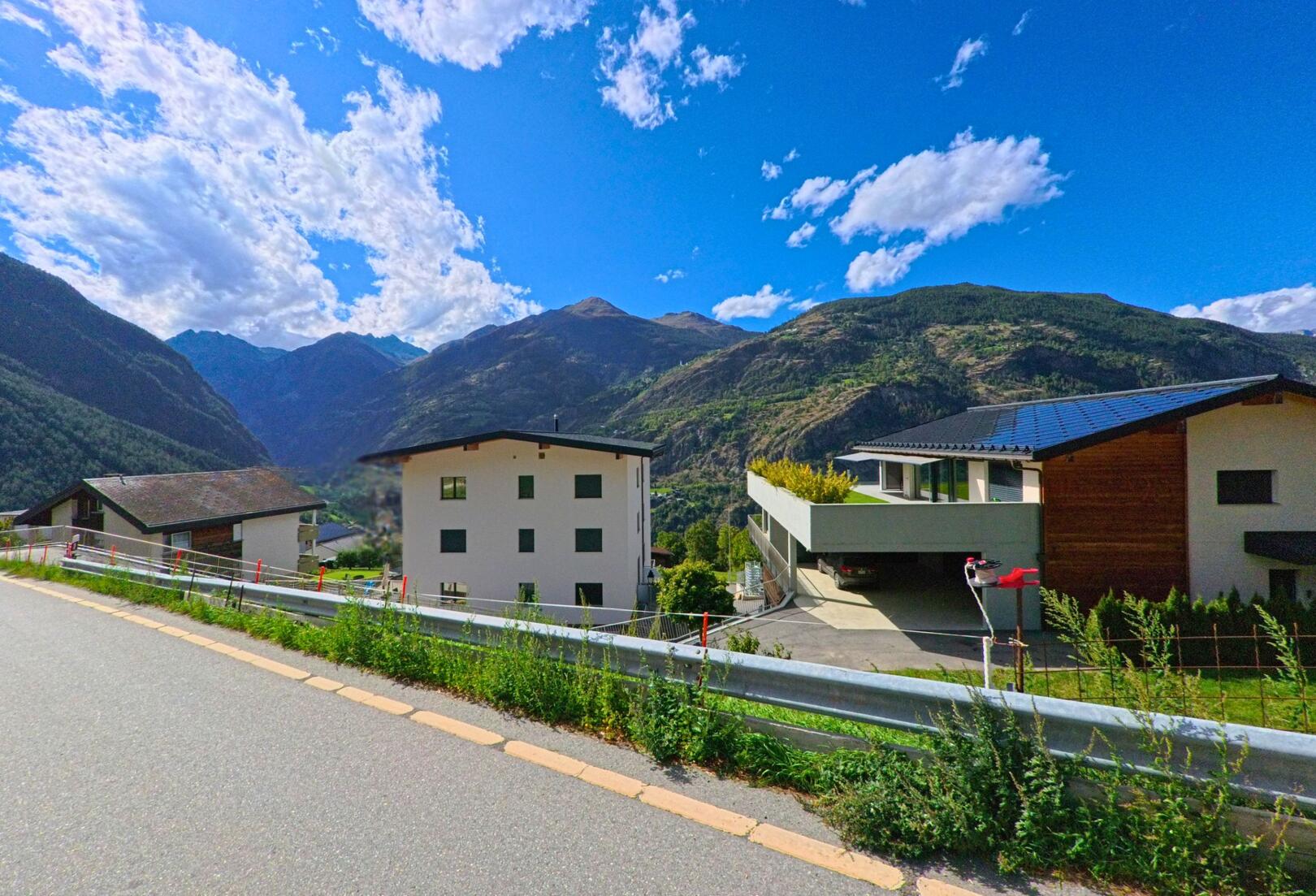 In Staldenried (3933, Kanton Wallis) stehen moderne Wohnhaeuser am Hang, umgeben von Bergen und blauem Himmel mit vereinzelten Wolken.