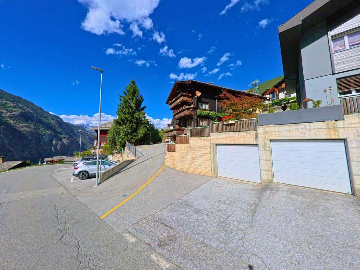 In Staldenried (3933, Kanton Wallis) fuehrt eine Strasse mit Parkplaetzen und Garagen an traditionellen Holzhäusern und modernen Bauten vorbei.