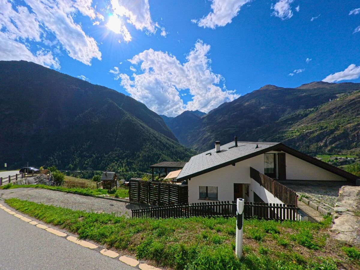 Ein weisses Wohnhaus mit Gartenzaun liegt in Staldenried (3933, Kanton Wallis) oberhalb des Tales mit Blick auf bewaldete Berge.