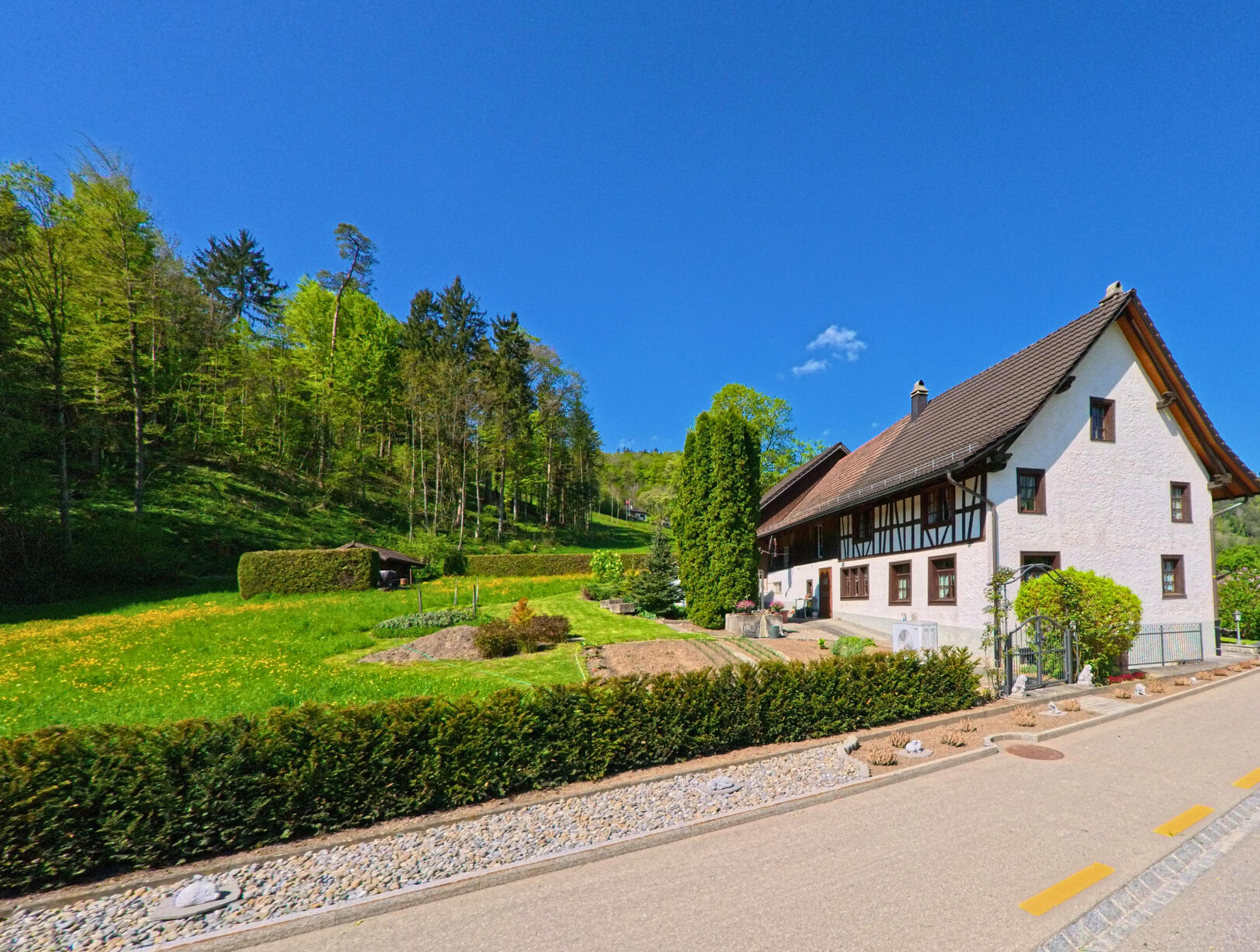 In Stallikon (8143, Kanton Zürich) steht ein traditionelles Bauernhaus mit gepflegtem Vorgarten am Waldrand unter klarem, blauem Himmel.