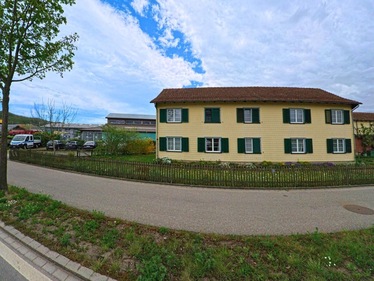 Ein langgestrecktes, gelb gestrichenes Wohnhaus mit grünen Fensterläden steht in Stammheim (8468, Kanton Zürich) hinter einem Holzzaun an einer ruhigen Strasse.