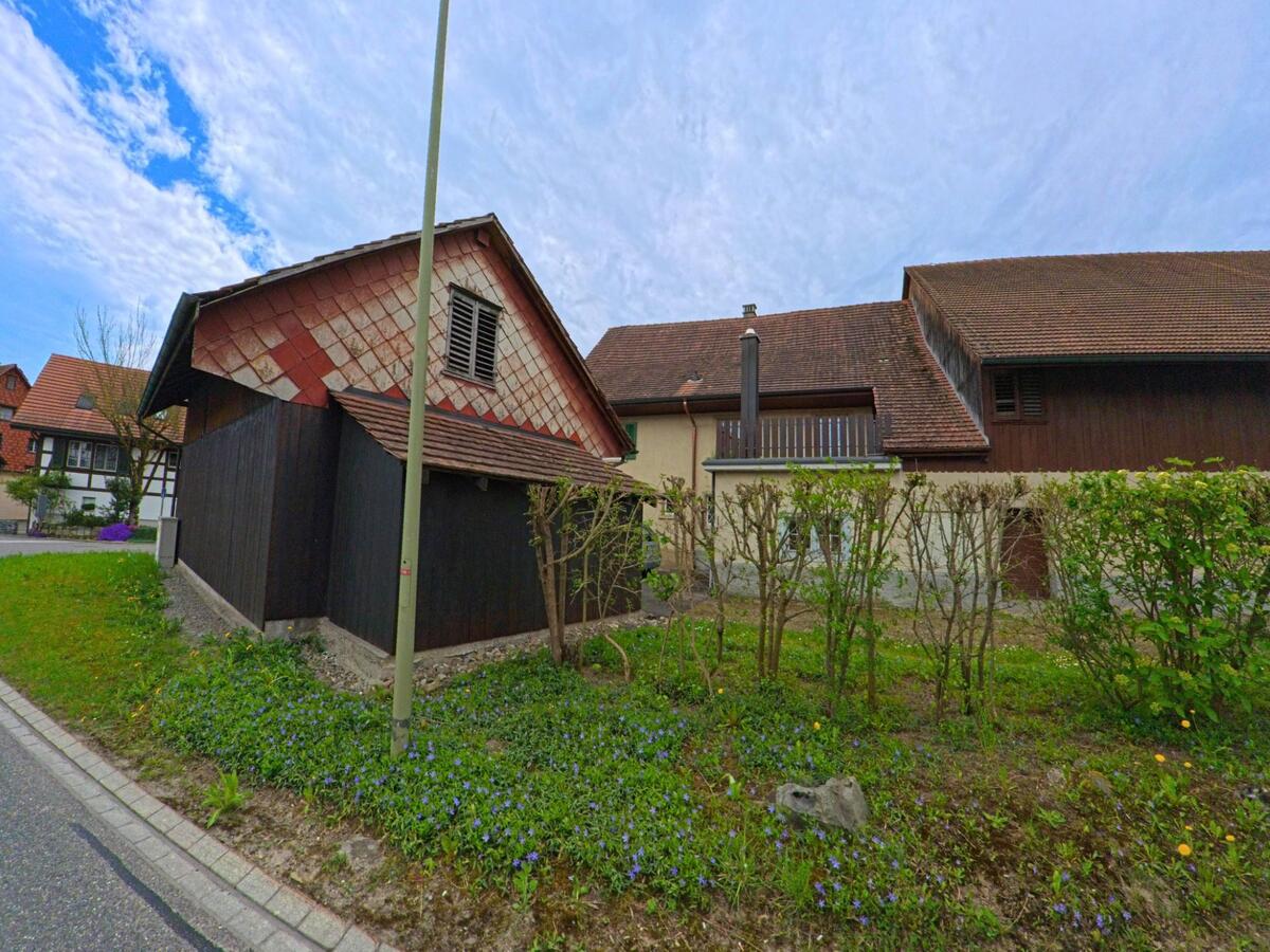 Ein kleiner Schuppen mit roten Dachschindeln steht in Stammheim (8468, Kanton Zürich) vor einem grösseren landwirtschaftlichen Gebäude mit Holzfassade.