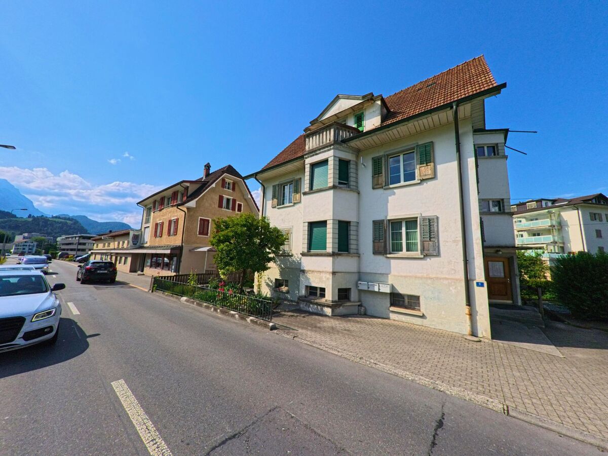 Entlang einer Hauptstrasse in Stans (6370, Kanton Nidwalden) reihen sich gepflegte Wohn- und Geschaeftshaeuser mit Fensterlaeden und Ladengeschaeften vor alpiner Kulisse aneinander.