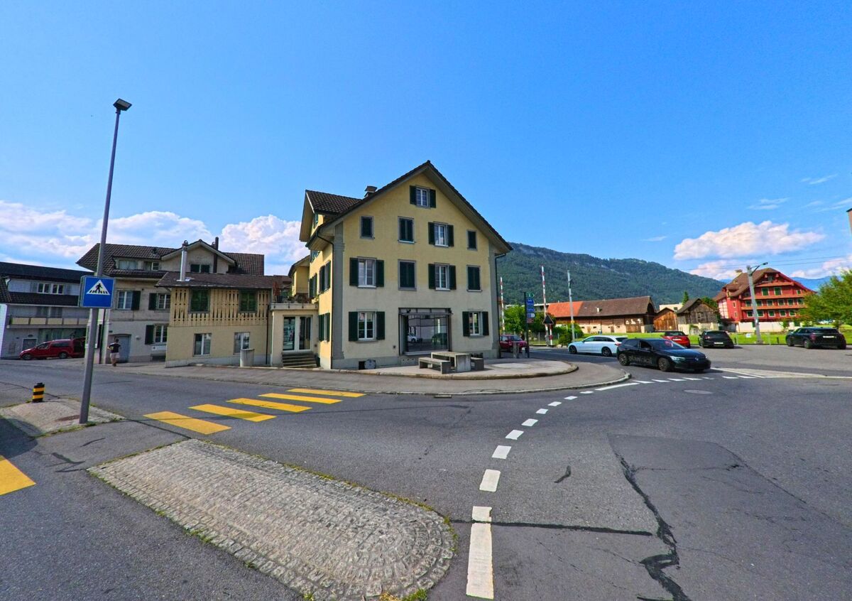 In Stans (PLZ 6370, Kanton Nidwalden) steht ein helles Eckhaus mit gruenen Fensterlaeden an einer Strassenkreuzung mit Fussgaengerstreifen und Blick auf die umliegenden Haeuser und Berghaenge.