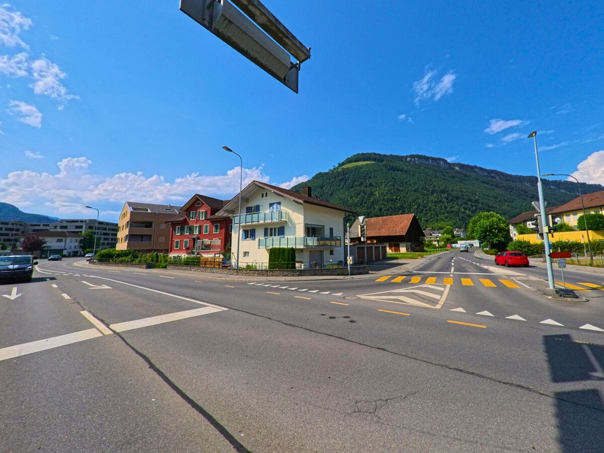 Mehrere Fahrstreifen und ein Fussgaengerstreifen kreuzen sich in Stans (PLZ 6370, Kanton Nidwalden) vor einer Reihe von Wohnhaeusern und einem bewaldeten Huegel im Hintergrund.