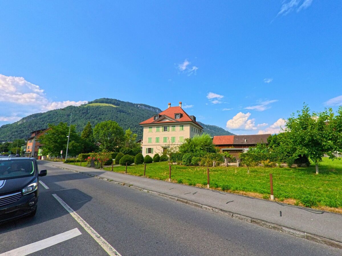 In Stans (PLZ 6370, Kanton Nidwalden) steht ein herrschaftliches Haus mit gruenen Fensterlaeden am Strassenrand, umgeben von gepflegten Gaerten und mit Blick auf einen bewaldeten Huegel.