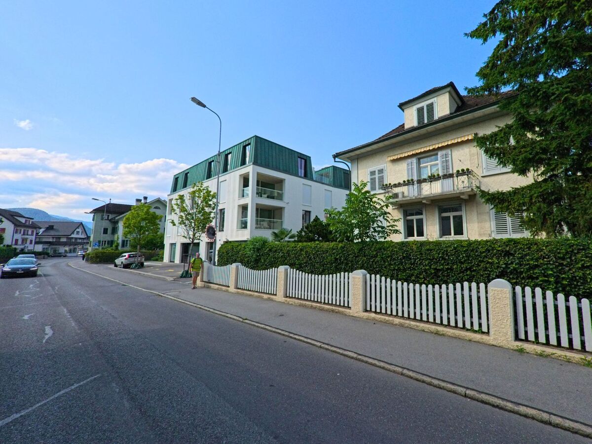 In einer ruhigen Wohnstrasse von Stans (6370, Kanton Nidwalden) stehen ein modernes Mehrfamilienhaus mit gruener Dacheindeckung und ein traditionelles Wohnhaus hinter einem weissen Zaun.