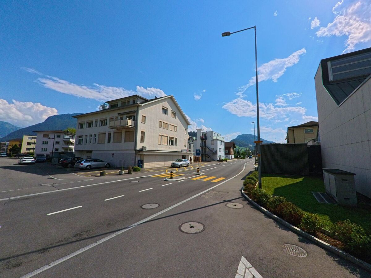 Entlang einer Durchgangsstrasse in Stansstad (6362, Kanton Nidwalden) stehen mehrstöckige Wohnhäuser mit Parkplätzen, im Hintergrund erheben sich bewaldete Hügel vor blauem Himmel.
