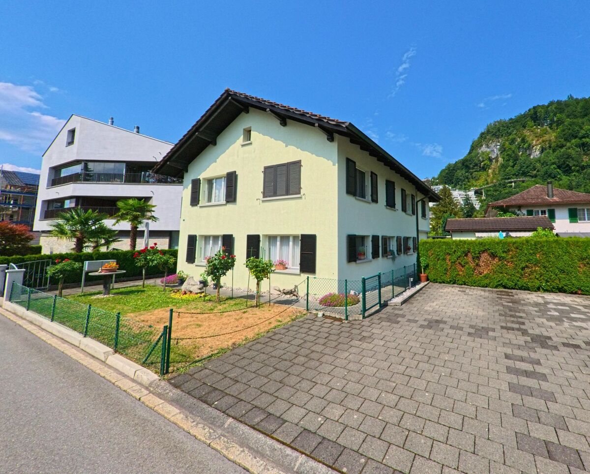 In Stansstad (6362, Kanton Nidwalden) steht ein hell gestrichenes Einfamilienhaus mit dunklen Fensterläden, kleinem Vorgarten und Blick auf bewaldete Hänge im Hintergrund.