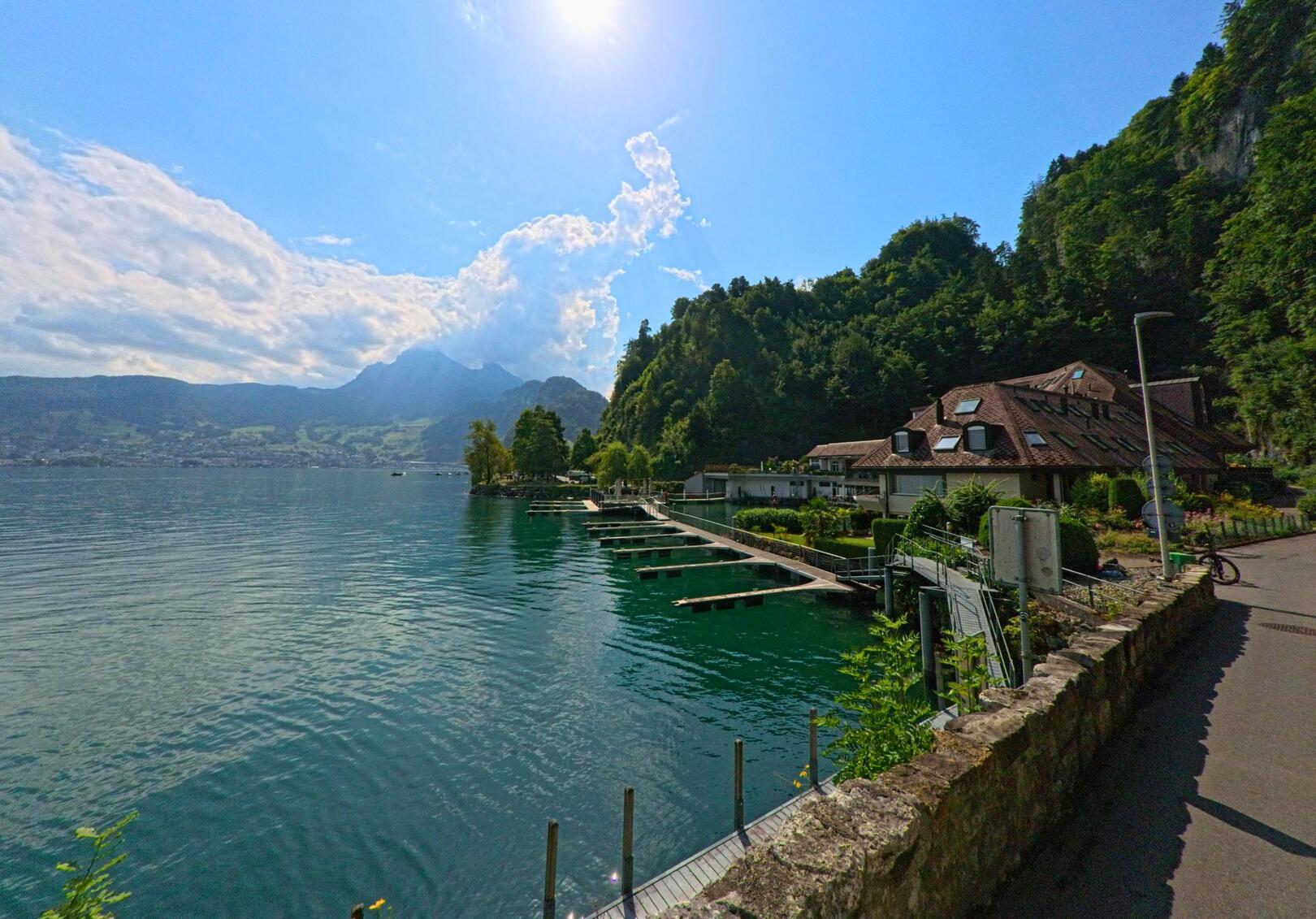 Direkt am Ufer des Vierwaldstaettersees in Stansstad (6362, Kanton Nidwalden) liegt eine kleine Hafenanlage mit Bootsstegen, umgeben von dicht bewaldeten Hängen und einem Haus mit markantem Dach.
